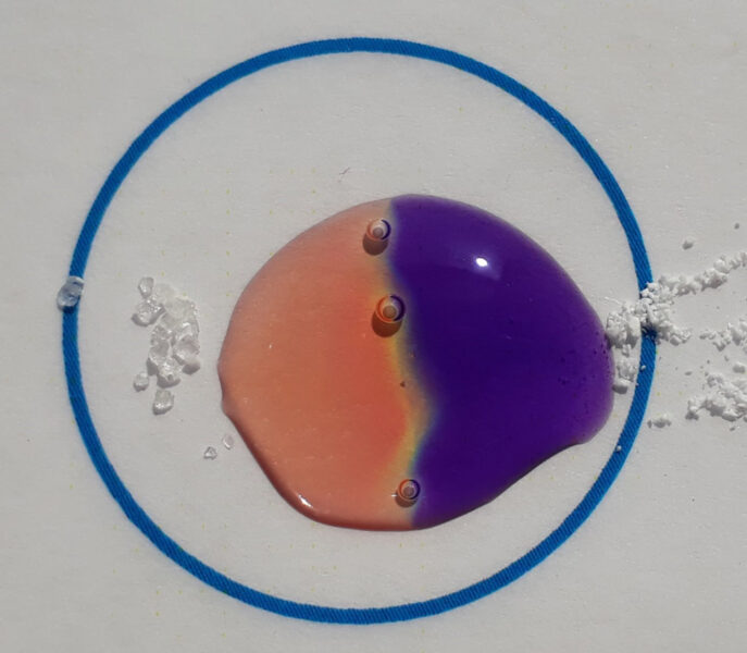 Al añadir dos sólidos a ambos lados de la gota una mitad se vuelve naranja y la otra, morada y se forma una línea amarilla con desprendimiento de burbujas donde se encuentran los dos colores.